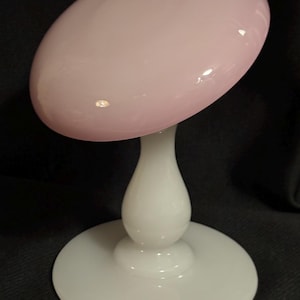 May include: A decorative glass object with a pink, oval-shaped top and a white, pedestal base. The pink top is smooth and glossy, while the base is a classic pedestal design. The object is likely a decorative piece.