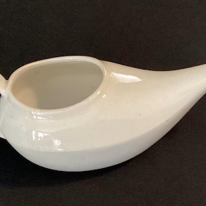 May include: A white ceramic neti pot with a curved spout and a handle. The pot has a wide opening and a smooth, glossy finish. It is designed for nasal irrigation and sits on a dark surface.