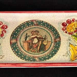 May include: Rectangular ceramic serving tray with a red rim and handles. The tray features a central circular design with a mosaic-style image and floral motifs on each side. The flowers are red and yellow with green leaves.