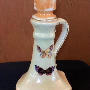 May include: Cream-colored ceramic candlestick holder with a handle and two butterfly decals. The top and base have an orange-brown trim. The candlestick holder is approximately 6 inches tall.