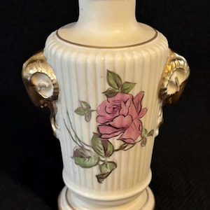 May include: A cream-colored ceramic vase with a pink rose and green leaves painted on the front. The vase has gold accents on the rim, base, and handles. The handles are gold and shaped like ram horns.