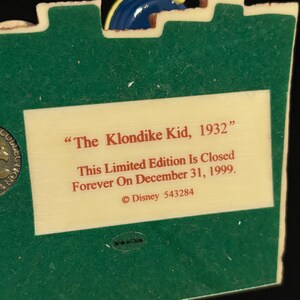 May include: A collectible figurine with a green base and a cream-colored plaque. The plaque reads "The Klondike Kid, 1932" and "This Limited Edition Is Closed Forever On December 31, 1999." A blue and yellow decorative element is at the top.