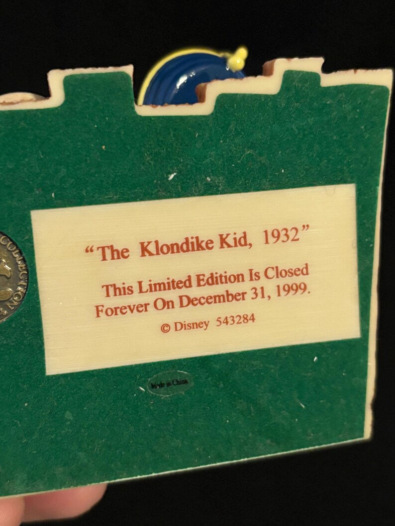 May include: A collectible figurine with a green base and a cream-colored plaque. The plaque reads "The Klondike Kid, 1932" and "This Limited Edition Is Closed Forever On December 31, 1999." A blue and yellow decorative element is at the top.