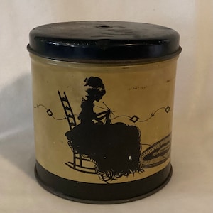 May include: A vintage yellow metal tin with a black lid. The tin features a black silhouette of a woman sitting in a rocking chair knitting. The woman is wearing a dress and a hat. The tin has a black rim around the top and bottom.