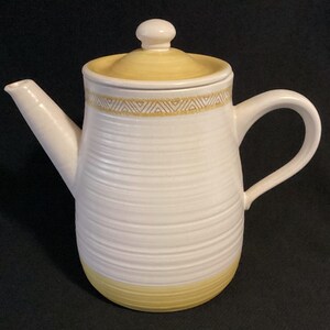 May include: A ceramic teapot featuring a cream-colored body with horizontal ribbed textures, a yellow base, and a matching yellow lid. A decorative band with a geometric triangle pattern in yellow and white encircles the upper section.