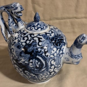 May include: A blue and white ceramic teapot with a dragon and floral design. The handle is shaped like a monkey, and the spout is a dragon's head. The lid has a leaf-shaped finial. The teapot is decorated with intricate patterns.