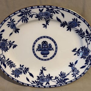 May include: Oval white serving platter with a blue floral design and gold trim. The platter features a central basket motif and a border of flowers and leaves. A classic piece for serving food.