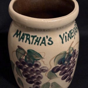 May include: A white ceramic jar with a brown rim and a green and purple painted grapevine design. The jar is labeled "Martha's Vineyard".