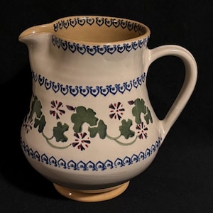 May include: A white ceramic pitcher with a curved handle and spout. The pitcher is decorated with green leaf and purple flower designs, and blue and white trim. The base and interior rim are tan.