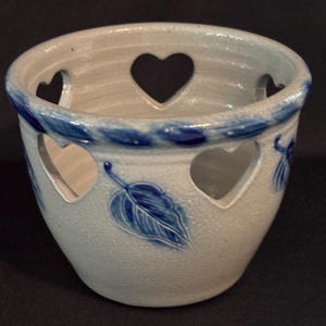 May include: A light gray ceramic bowl featuring a series of cut-out heart shapes around the upper rim. The exterior is decorated with embossed blue leaf patterns and a textured blue rope-like border encircling the top edge of the vessel.