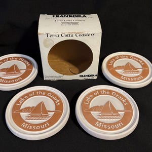 May include: A set of four white terra cotta coasters with a brown and white graphic of a sailboat on the water, with the words "Lake of the Ozarks Missouri" around the edge. The coasters are next to a box that says "Terra Cotta Coasters".