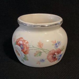 May include: A small, round, off-white ceramic jar with a silver rim. The jar is decorated with a floral pattern featuring red and pink poppies, blue flowers, and green leaves. The jar sits on a black surface.