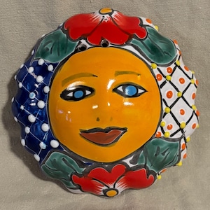 May include: A round ceramic wall plaque featuring a central yellow sun face with blue eyes and a dark mouth. The border is decorated with red flowers, green leaves, and two distinct patterned sections: one dark blue with white dots and one white with a grid.