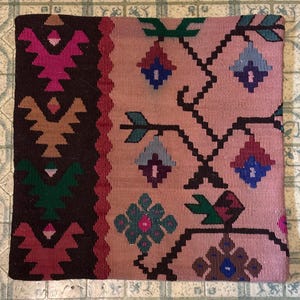 May include: A square, woven pillow with a detailed, geometric pattern. The pillow features a central field of muted rose with dark brown, green, and blue accents. The border is a darker brown with repeating geometric shapes.