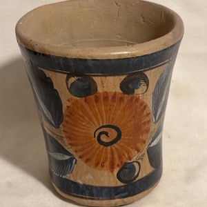 May include: Hand-painted ceramic cup with a beige base, adorned with a large orange floral design and dark blue accents. The cup features a dark blue band near the rim and is likely handmade.