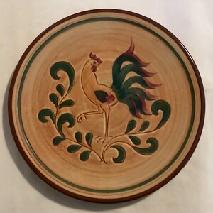 May include: Round ceramic plate with a rooster design. The plate features a tan background with a green and brown border. A yellow rooster with green and red tail feathers is in the center, surrounded by green leaf accents.