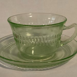 May include: A light green glass teacup and saucer set. The cup has a handle and a textured, rectangular pattern around the body. The saucer also has a textured design. The set is made of transparent glass.