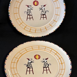 May include: Two white ceramic plates with a yellow grid pattern and a colorful dot border. Each plate features a hand-painted design of two dogs wearing red hats, sitting on stools and playing with a yellow ball.