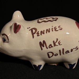 May include: White ceramic piggy bank with brown lettering that reads "Pennies Make Dollars." The pig has red and blue accents, including heart-shaped ears and blue hooves. A classic savings item.