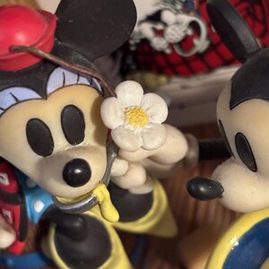 May include: A figurine featuring Mickey and Minnie Mouse. Minnie Mouse is holding a white flower with a yellow center. Mickey Mouse is wearing a yellow and blue outfit. Minnie Mouse is wearing a red hat and a blue and red outfit.