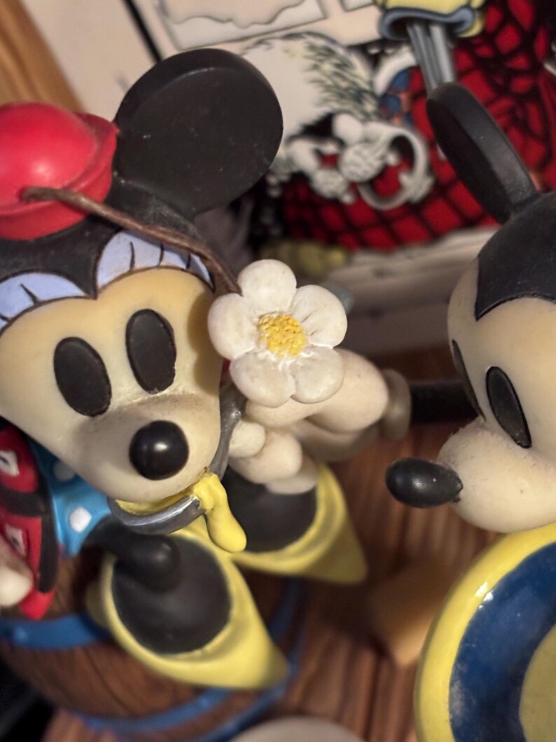 May include: A figurine featuring Mickey and Minnie Mouse. Minnie Mouse is holding a white flower with a yellow center. Mickey Mouse is wearing a yellow and blue outfit. Minnie Mouse is wearing a red hat and a blue and red outfit.