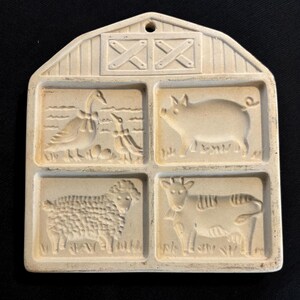 Puede incluir: Molde de cer&aacute;mica en forma de granero, de color blanquecino, con cuatro im&aacute;genes de animales en relieve: un ganso con huevos, un cerdo, una oveja y una vaca. El tejado del granero tiene dos cruces decorativas y un agujero para colgar.