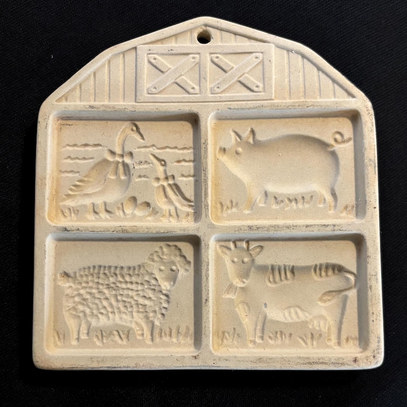 Puede incluir: Molde de cer&aacute;mica en forma de granero, de color blanquecino, con cuatro im&aacute;genes de animales en relieve: un ganso con huevos, un cerdo, una oveja y una vaca. El tejado del granero tiene dos cruces decorativas y un agujero para colgar.