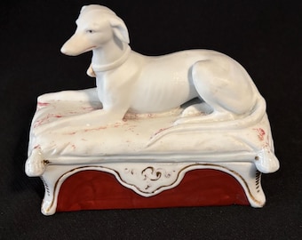 Caja de baratijas antigua de porcelana bizcochada con forma de perro galgo sobre un sofá.