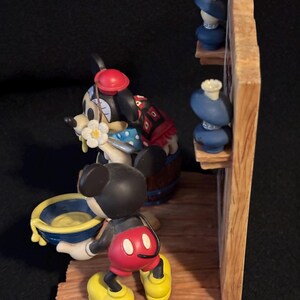 May include: A detailed figurine featuring Mickey Mouse and Minnie Mouse. Mickey is holding a blue and yellow bowl. Minnie is wearing a red hat and a blue polka dot dress. The scene includes a wooden structure with blue decorative items.