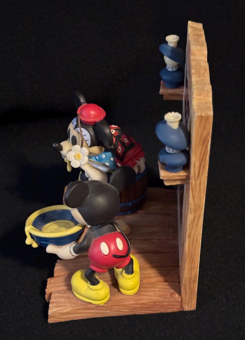 May include: A detailed figurine featuring Mickey Mouse and Minnie Mouse. Mickey is holding a blue and yellow bowl. Minnie is wearing a red hat and a blue polka dot dress. The scene includes a wooden structure with blue decorative items.