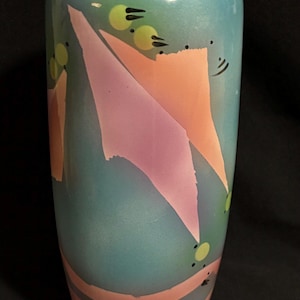 May include: A tall, cylindrical vase with a teal background and abstract geometric shapes in pink and orange. The vase has a smooth finish and is decorated with small yellow circles and black accents.