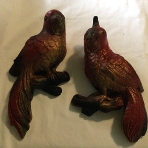 May include: Two red and gold painted wooden parrot figurines. The parrots are perched on branches and have detailed feathers.