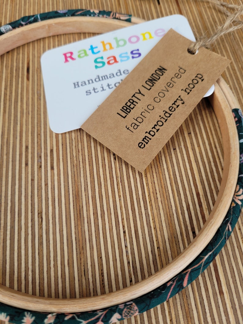 Peut inclure: Un cerceau de broderie en bois avec un rev&ecirc;tement en tissu pr&eacute;sentant un motif floral dans des tons de vert et de rose. Le cerceau est &eacute;tiquet&eacute; "Liberty London tissu recouvert de broderie" et "Rathbone Sass Couture faite main".