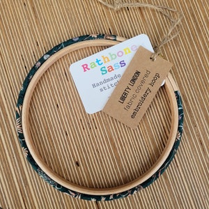 Peut inclure: Un cercle &agrave; broder recouvert de tissu floral vert et blanc avec une &eacute;tiquette brune indiquant "Liberty London tissu recouvert cercle &agrave; broder" et une &eacute;tiquette blanche indiquant "Rathbone Sass Couture faite main".