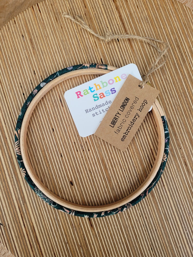 Peut inclure: Un cercle &agrave; broder recouvert de tissu floral vert et blanc avec une &eacute;tiquette brune indiquant "Liberty London tissu recouvert cercle &agrave; broder" et une &eacute;tiquette blanche indiquant "Rathbone Sass Couture faite main".