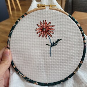 Peut inclure: Gros plan d'un projet de broderie au point de croix termin&eacute; repr&eacute;sentant une seule fleur rouge avec des feuilles vertes dans un cercle de broderie en bois rond avec une bordure en tissu floral.