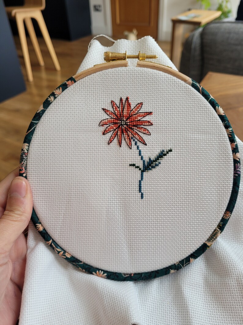 Peut inclure: Gros plan d'un projet de broderie au point de croix termin&eacute; repr&eacute;sentant une seule fleur rouge avec des feuilles vertes dans un cercle de broderie en bois rond avec une bordure en tissu floral.