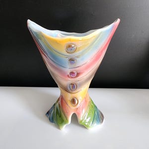 以下が含まれることがあります： ユニークな形と虹色のアクセントが施されたカラフルなセラミックの花瓶。花瓶は、黄色、青、赤、緑など、虹色の色で飾られています。開口部が広く、底が狭い.