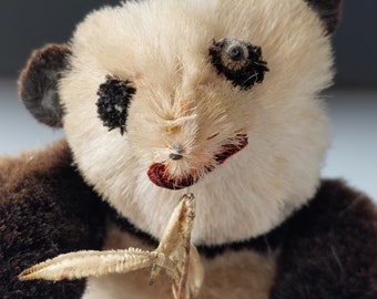 Puede incluir: Un juguete de peluche vintage de panda con pelaje negro alrededor de los ojos y las orejas, sosteniendo una rama marrón en sus patas. La piel del panda es blanca y tiene un aspecto desgastado y deteriorado.