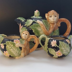 Puede incluir: Un juego de té negro con diseño de mono. La tetera tiene un mono agarrado a una hoja, la jarra de crema tiene un mono agarrado a una hoja y el azucarero tiene un mono agarrado a una hoja. El juego está decorado con hojas verdes y flores rosas.