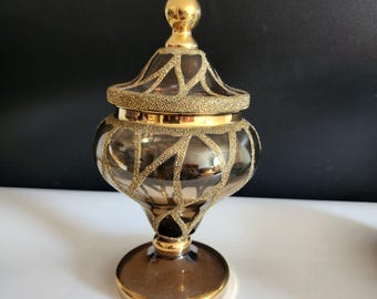 May include: A decorative glass jar with a lid, featuring a gold finial and trim. The jar has a brown tint and is adorned with a gold geometric pattern. The base is also gold, creating an elegant, vintage aesthetic.
