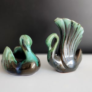 Może przedstawiać: Dwie ceramiczne figurki łabędzi w zielonym i brązowym szkliwie. Łabędzie siedzą z zakrzywionymi szyjami.