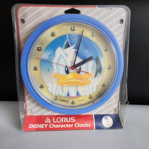 May include: A blue and yellow wall clock featuring a cartoon image of Donald Duck with the text "LORUS QUARTZ" on the face. The clock is packaged in a clear plastic box with the text "LORUS DISNEY Character Clocks" on the front.