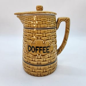 May include: A tan ceramic coffee pot with a basket weave design. The pot has a lid and a handle. The word "COFFEE" is displayed in black script on the front of the pot. The pot is approximately 8 inches tall.