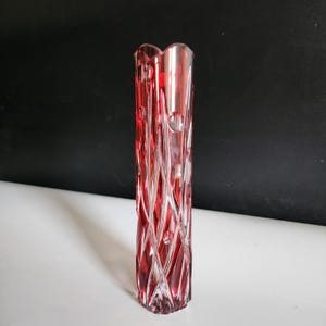 May include: A tall, red glass vase with a geometric pattern. The vase has a square base and a flared top with a heart-shaped design. The red colour is translucent, allowing light to pass through.
