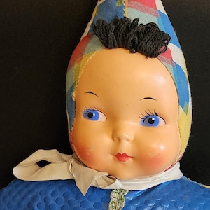 May include: A vintage doll with a blue and white patterned dress, a yellow and red patterned hat, and blue eyes. The doll has a painted face and black yarn hair.