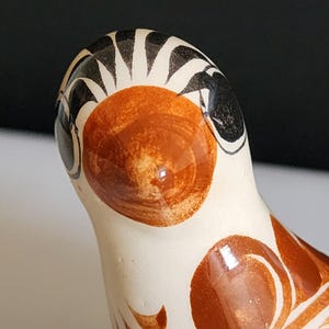 Peut inclure: Gros plan sur une figurine d'oiseau en céramique. L'oiseau a une couleur de base blanche avec des motifs décoratifs marron et noirs. La tête de l'oiseau présente une grande zone brun-orangé ronde et les yeux sont noirs. Le design est peint à la main.
