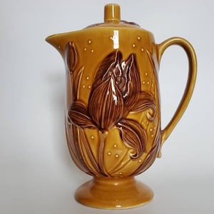 May include: A brown and gold ceramic pitcher with a floral design. The pitcher has a lid and a handle. The floral design is of a brown flower with a yellow background.