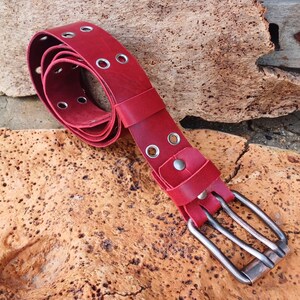 May include: A red leather belt with silver-colored grommets and a rectangular silver buckle. The belt is shown coiled on a textured, light brown surface.