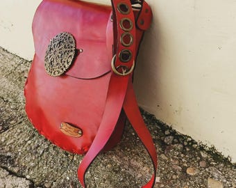May include: A red leather satchel with a decorative metal clasp and a long strap. The bag has a flap closure and a small wooden tag. The strap is adjustable with metal eyelets and a buckle. The bag is leaning against a wall.